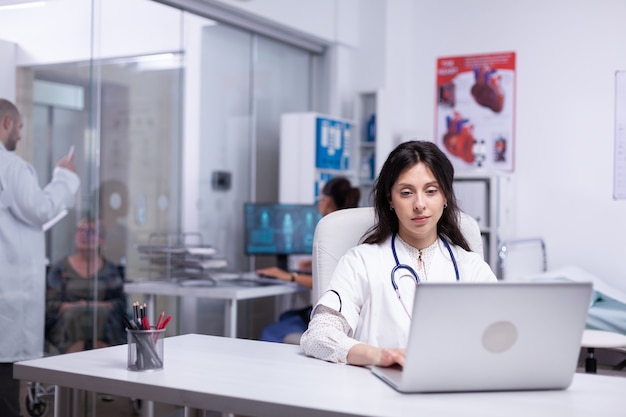 Professional doctor in white coat working on laptop in modern hospital office room, therapist typing on computer consult patient online, makes research, analysing results information from internet