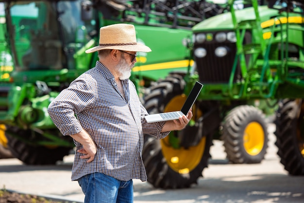 Professional farmer with a modern tractor at work with laptop. Looks confident, bright summer colors, sunshine. Agriculture, exhibition, machinery, plant production. Senior man near his machine.