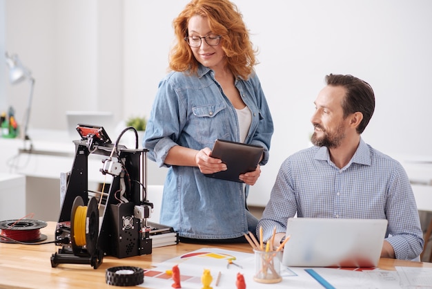 Professional female 3d designer holding a tablet and looking at the 3d printer while working with her colleague in the office