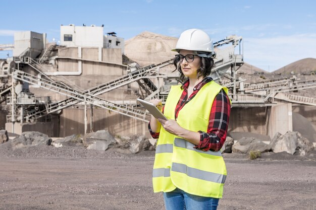 Architetto professionista femminile in elmetto protettivo e panciotto controllo progetto su tablet mentre si trovava vicino all'impianto industriale del cantiere