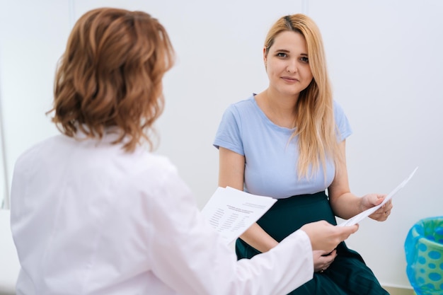 Professional female doctor transmits to young pregnant woman with blonde hair the results and reports of tests and conducted looking at camera Concept of happy and healthy childbearing