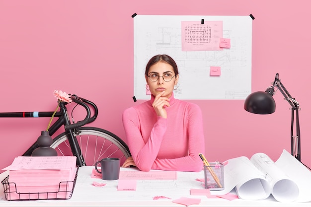 Professional female engineer thinks over ideas for building project has thoughtful expression wears round spectacles and turtleneck poses in coworking space against pink wall blueprint behind