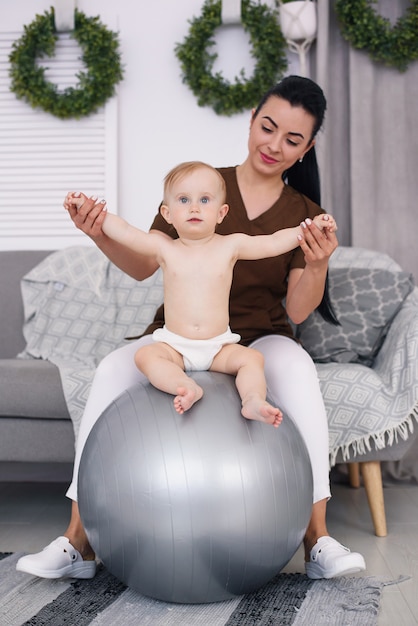 A professional female masseuse with happy baby doing exercises with fitness ball at medical room. Healthcare and medical concept.