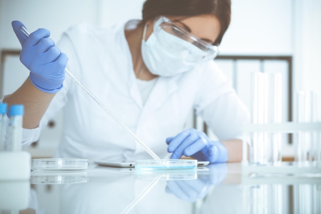 Professional female scientist in protective eyeglasses and mask making experiment with reagents in laboratory. Coronavirus pandemic stop. Medicine and research concept.