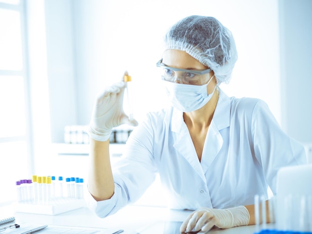 Professional female scientist in protective eyeglasses researching tube with reagents in laboratory toned in blue. Concepts of medicine and science researching.