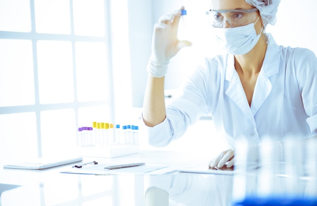 Professional female scientist in protective eyeglasses researching tube with reagents in sunny laboratory toned in blue. Medicine and science researching.