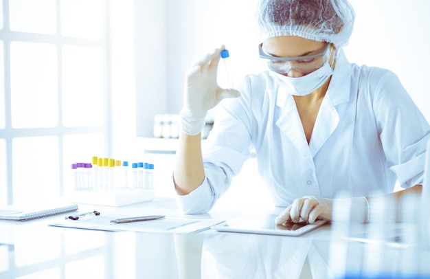 Professional female scientist in protective eyeglasses researching tube with reagents in sunny laboratory toned in blue Medicine and science researching