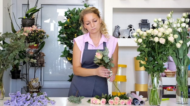 Профессиональный флорист за работой в цветочном магазине. Женщина в униформе стоит возле стола и делает букет