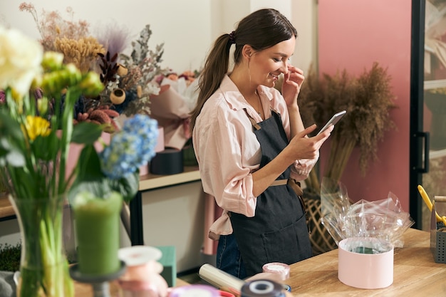 Professional florist young woman cominucating with clients via smartphone at flower shop
