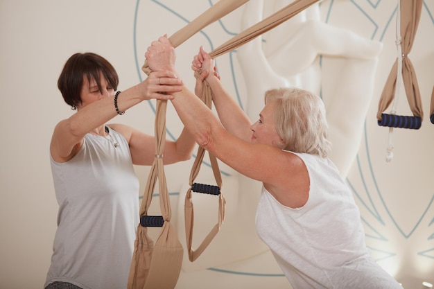 Professional fly yoga instructor helping her elderly client exercising on aerial yoga hammock