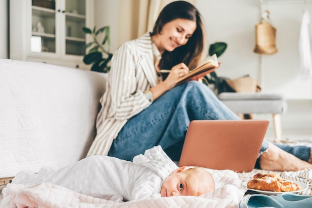 Tipi di liberi professionisti professionisti su laptop grigio seduto su un tappeto con vestiti alimentari e bambino piccolo che dorme su una coperta bianca a casa.