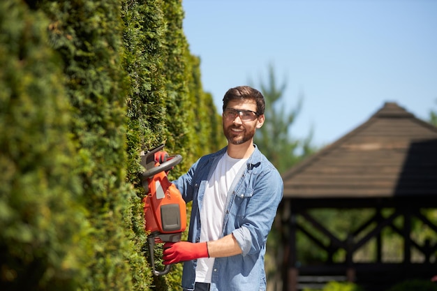 professional gardener trimming shaping evergreen thuja hedge with modern electric trimmer at summer