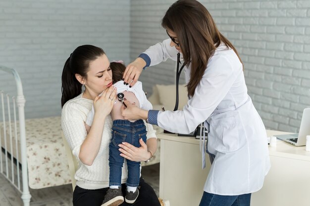 Профессиональный врач-педиатр общего профиля в белом халате при помощи стетоскопа слушает звук легких и сердца ребенка.