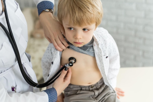 Профессиональный врач-педиатр общего профиля в белом халате при помощи стетоскопа слушает звук легких и сердца ребенка.