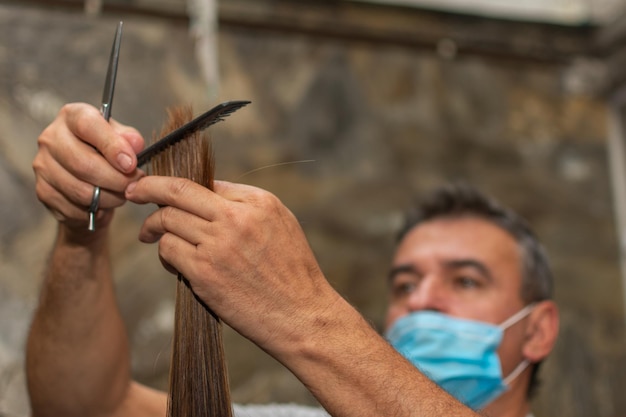 Professional hairdresser Focus on hand holding a lock of hair while his face covered by a medical face mask is blurred in the background