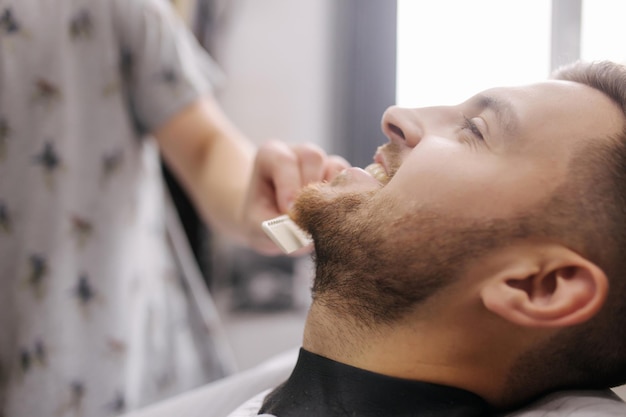 Professional hairdresser uses a hair clipper for fringing beard for a handsome man in barbershop side view