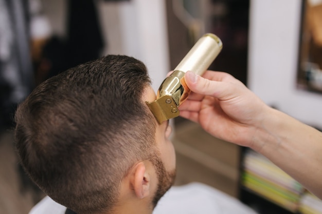 Professional hairdresser uses a hair clipper for fringing hair for a handsome bearder man. Barbershop. Closeup.