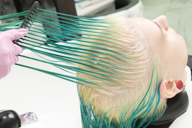 Professional hairstylist in protective glove holds of hair in hand and brushes long green and bleached hair of client, while washing head in shower in special sink of beauty salon.