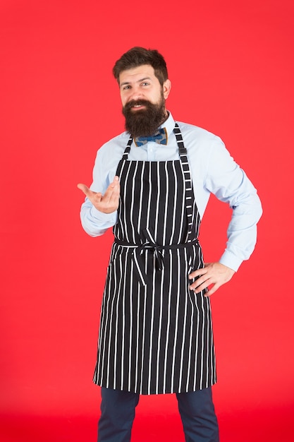 주방 전문. 수염을 가진 남자 쿡 힙스터 앞치마. Hipster 요리사는 빨간색 배경을 요리합니다. 수염 난 남자 요리사 요리입니다. 힙스터 요리 집이나 레스토랑. 현대적인 카페 컨셉입니다. 현대식 요리.