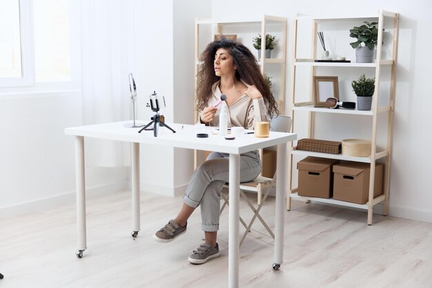 Professional makeup artist tanned handsome curly latin beauty blogger in linen shirt tells how to sculpt body in home white interior copy space influencer record video review blog using smartphone