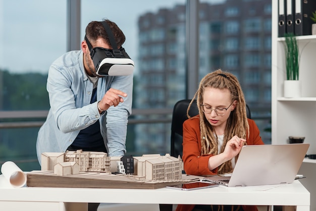 Professional male architect in augmented reality goggles working with mock up of building and woman