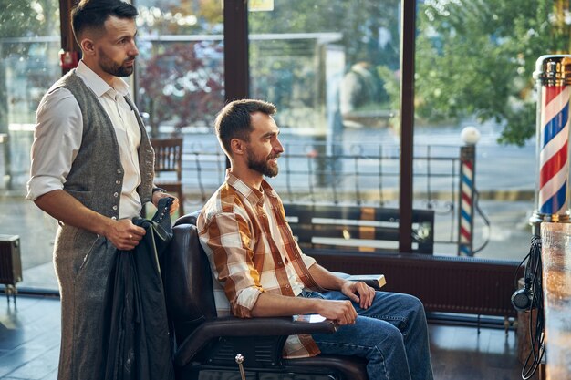 Professional male barber standing behind an armchair with a client at a modern barbershop