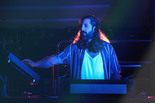 Professional male in casual wear playing song at DJ setup with audio mixer and CDJ while performing music in dark nightclub