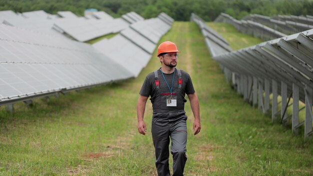 保護ヘルメットのプロの男性エンジニアは、生態学的な太陽光発電所の建設を歩きます。電気、エコロジー、テクノロジーの概念。ソーラーパネルの農場。