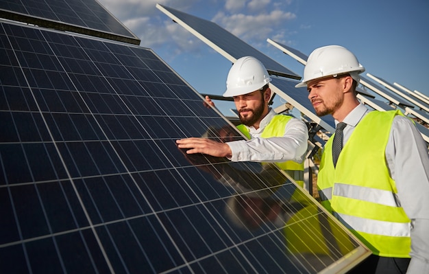 Professional male engineers in hardhats and waistcoats inspecting photovoltaic panels for alternative energy on solar power station