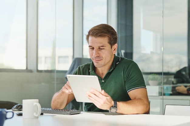 professional male entrepreneur in green polo sitting at desk with keyboard and browsing tablet while working on project in creative workspace