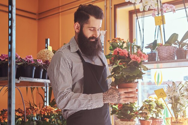 制服を着た彼の手にひげと入れ墨を持つプロの男性花屋は、フラワーショップで花の花束で鍋を保持します。