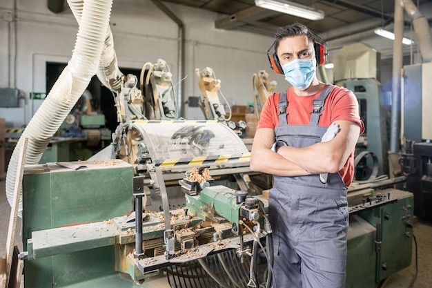 Professional male master in protective uniform and medical mask folding hands while standing near wood turning machine