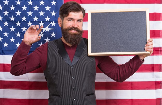 Professional man Man teacher giving lesson on american flag background Bearded man holding scissors and blackboard in school Man barber providing knowledge and skills of cutting hair copy space