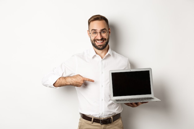 Professional manager showing webpage on laptop screen, pointing at computer, standing over white background.