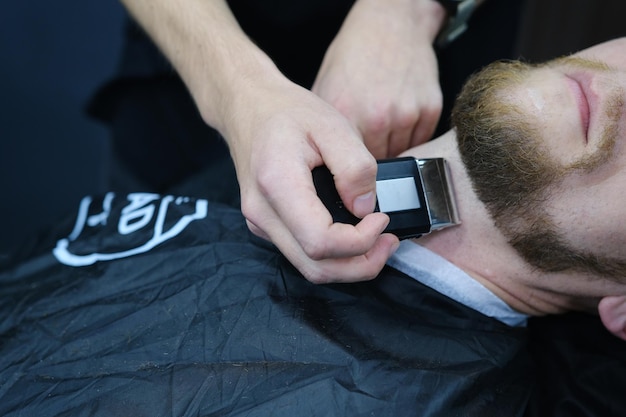 プロのマスター理髪師は、電気トリマーでクライアントのひげを剃ります 理髪店で男性のひげのヘアカット 理髪店の男性広告と理髪店のコンセプト