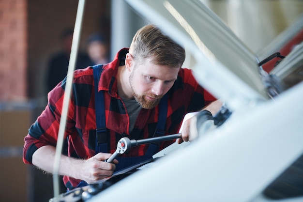 Professional master of car repair using one of handtools while bending over open engine in workshop