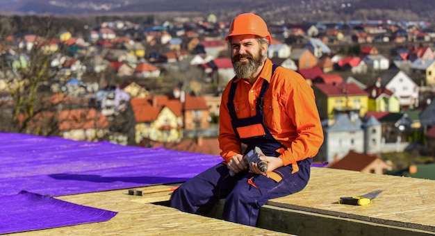 Photo professional master repair roof. flat roof installation. roofing felt. roofer worker in protective work wear. new roof under construction residential building. builder use drill. foreman planning.