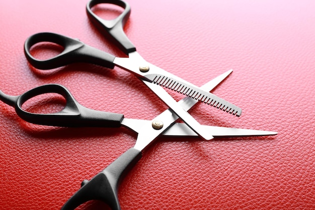 Professional metal scissors with black handles lying crossed on red leather surface close up