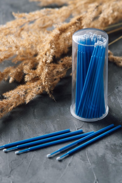 Профессиональные микро щетки с голубой палкой, лежащие в контейнере и оставленные на сером столе рядом с ветвями бежевой травы пампы Инструмент для процедуры удлинения ресниц с использованием в качестве аппликатора Вертикальный