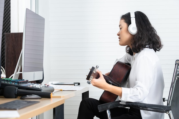 전문 음악가 젊은 아시아 여성 보컬인 Wearing Headphones는 전문 스튜디오에서 소리를 녹음하고 편집하기 위해 소프트웨어 컴퓨터 모니터의 노래를 기타로 녹음합니다.