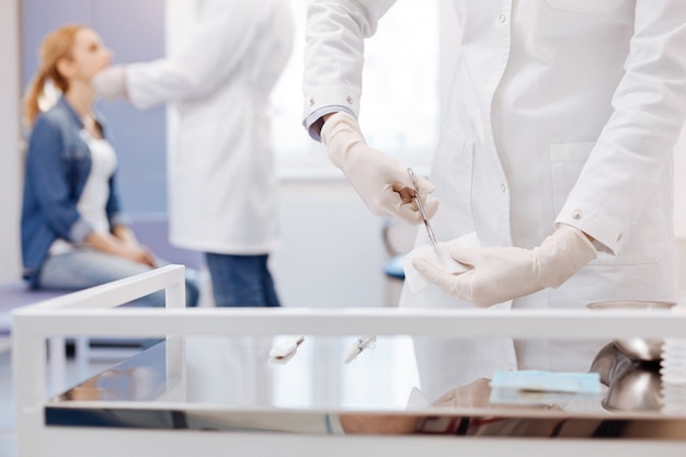 Professional nice experienced doctor standing near the table and wearing disposable gloves while cutting a piece of bandage