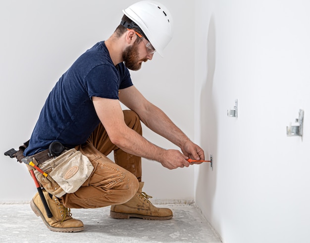 Professional in overalls with an electrician's tool on the white wall. Home repair and electrical installation concept.