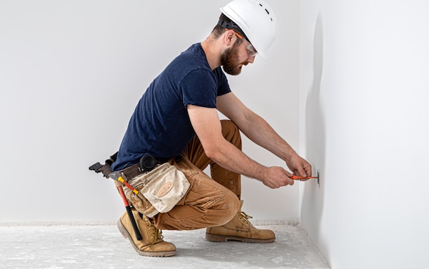 Professional in overalls with an electrician's tool on the white wall. Home repair and electrical installation concept.