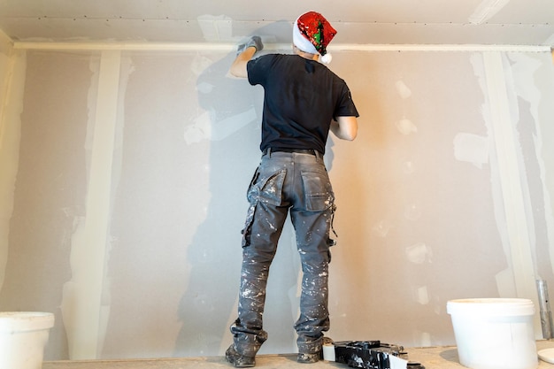 Professional painter in Santa hat applying plaster on wall for home renovation