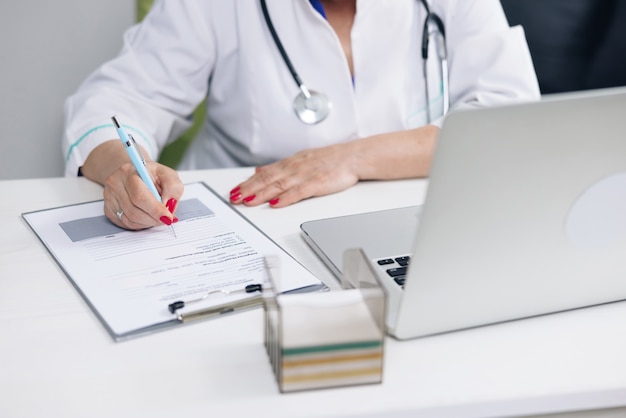 Professional physician wearing white coat writing notes using laptop at desk