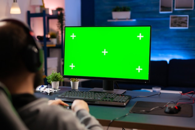 Photo professional player streaming videogames with green screen mock-up computer display in gaming home studio. gamer using pc with chroma key isolated desktop streaming shooter games wearing headset