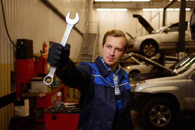 Professional portrait of a young Caucasian auto mechanic in uniform holding a wrench while standing at his workplace in a car service. Car repair and maintenance concept.