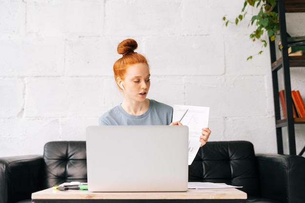 Professional redhead young businesswoman wearing wireless\
earphones is communicating by video call on laptop computer with\
colleagues lady picks up stack of paperwork documents and reports\
to boss