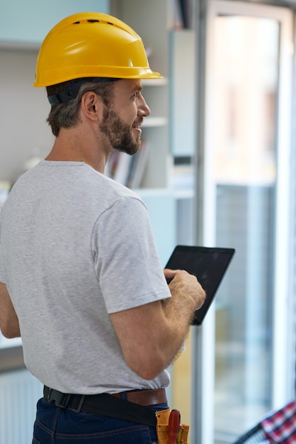 屋内に立っている間、デジタルタブレットを使用して脇を見てヘルメットをかぶったプロの修理工