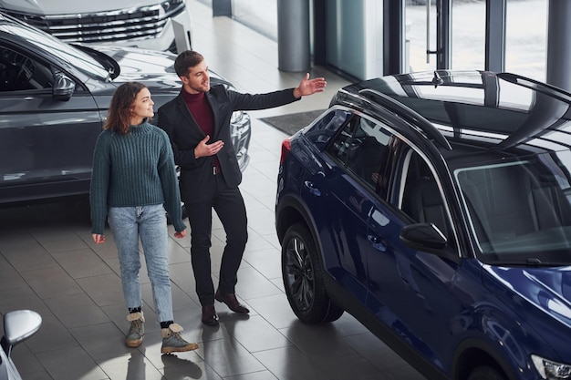 Professional salesman assisting young girl by choosing new modern automobile indoors.